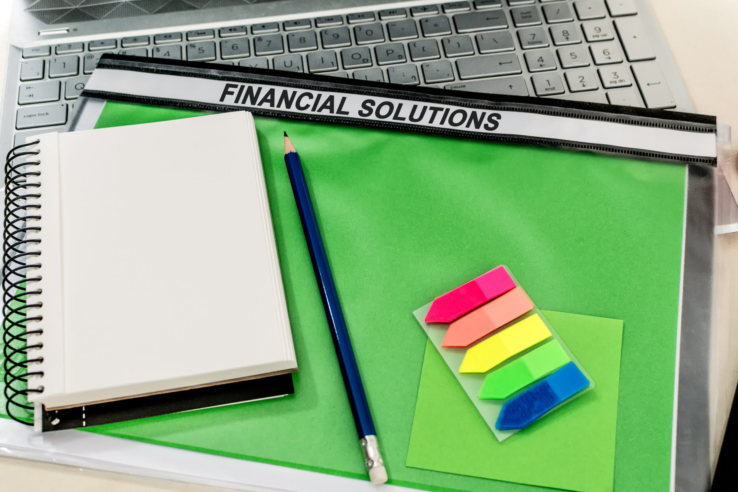 A notepad with a pencil on a green folder labelled "FINANCIAL SOLUTIONS" on a laptop in the office